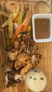 Grilled chicken pieces served on a wooden board with a side of mixed sautéed vegetables, a small portion of fried rice, and a square bowl of brown sauce.
