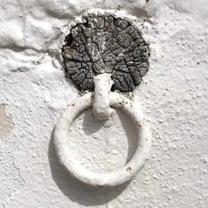 White painted metal ring attached to an exterior wall. The wall paint is chipped around the ring, revealing a weathered, cracked texture underneath.