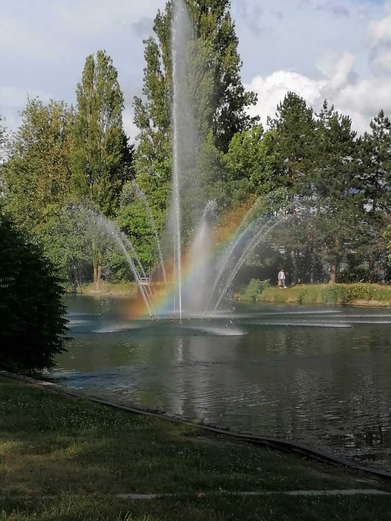 A picturesque scene of a pond with fountains spraying water into the air, creating a vibrant rainbow effect. Trees surround the pond, and a person can be seen walking on a path in the background.