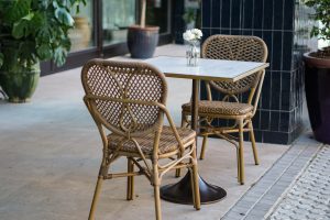 A small outdoor café setting with two wicker chairs at a square marble-topped table, with a single clear vase holding two white roses.