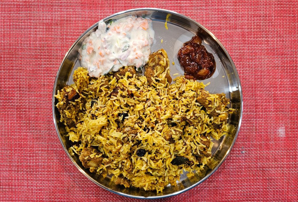 A plate of home-made chicken dum biriyani served with dates pickles and raita on a red table.