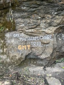 Rock surface with multiple graffiti inscriptions, including prominent white and yellow text reading "GREAT MACHHAPUCHHRE TRAIL". There are also various smaller carvings and writings scattered across the rock. Moss and plants are visible on the rock and surrounding area.