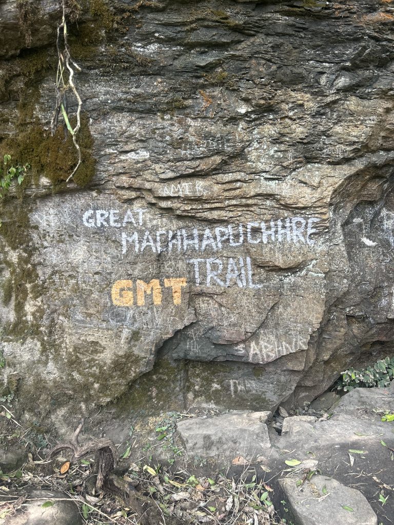 Rock surface with multiple graffiti inscriptions, including prominent white and yellow text reading “GREAT MACHHAPUCHHRE TRAIL”. There are also various smaller carvings and writings scattered across the rock. Moss and plants are visible on the rock and surrounding area.