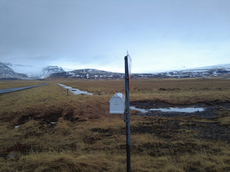 A rural landscape of Iceland featuring a mailbox labeled “U.S. Mail” on a post beside a gravel road. In the background, there are rolling hills and snow-capped mountains under a cloudy sky. The ground is covered in dry grass with patches of snow and puddles.