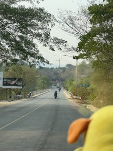 View larger photo: A view from inside a vehicle approaching a bridge called "PUENTE RíO COCO," with two motorcyclists ahead on the road, green trees on the sides, under a clear sky, and a power line running parallel to the road. The foreground shows a blurred part of the vehicle's interior, with a hint of a yellow object, possibly a toy or accessory.