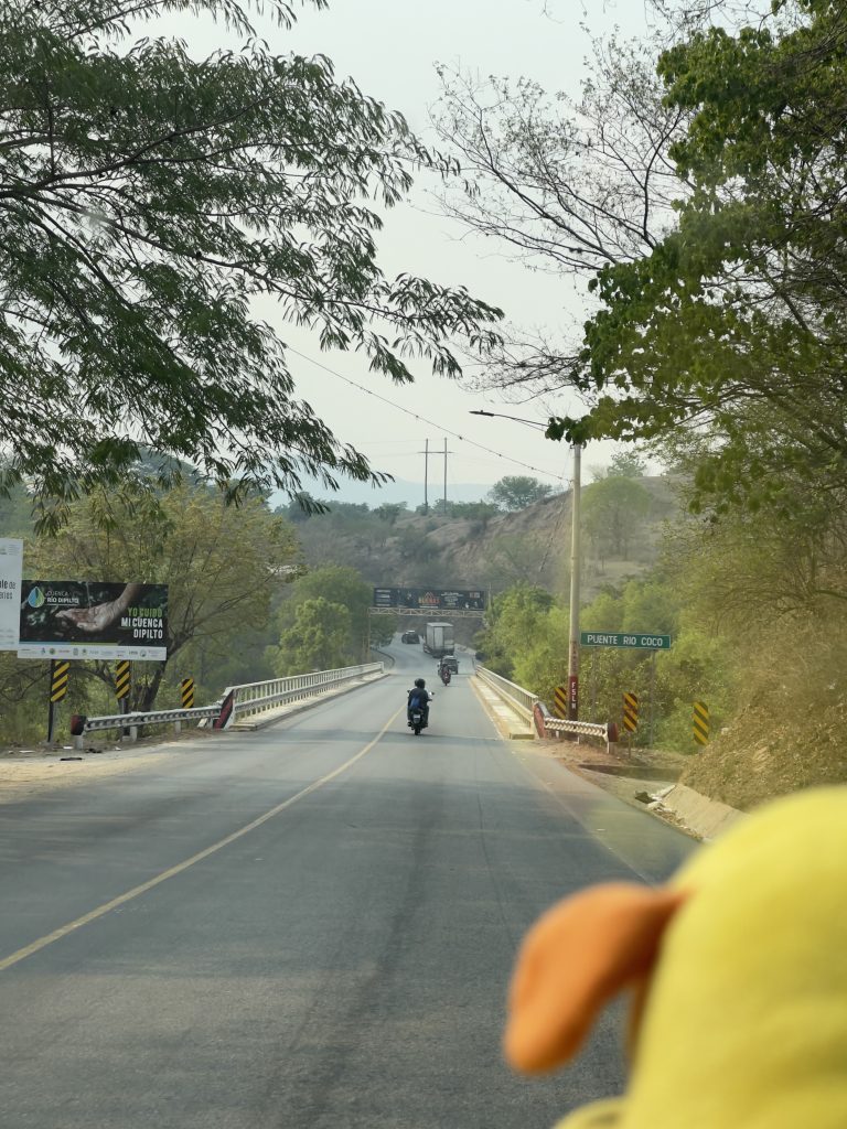 A view from inside a vehicle approaching a bridge called “PUENTE RíO COCO,” with two motorcyclists ahead on the road, green trees on the sides, under a clear sky, and a power line running parallel to the road. The foreground shows a blurred part of the vehicle’s interior, with a hint of a yellow object, possibly a toy or accessory.