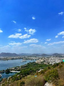 The photo captures a stunning aerial view of a city nestled around a large lake, Udaipur in India. The lake is dotted with small boats and features islands with structures. Surrounding the lake are lush green areas, historic buildings, and a dense cluster of white-washed houses.