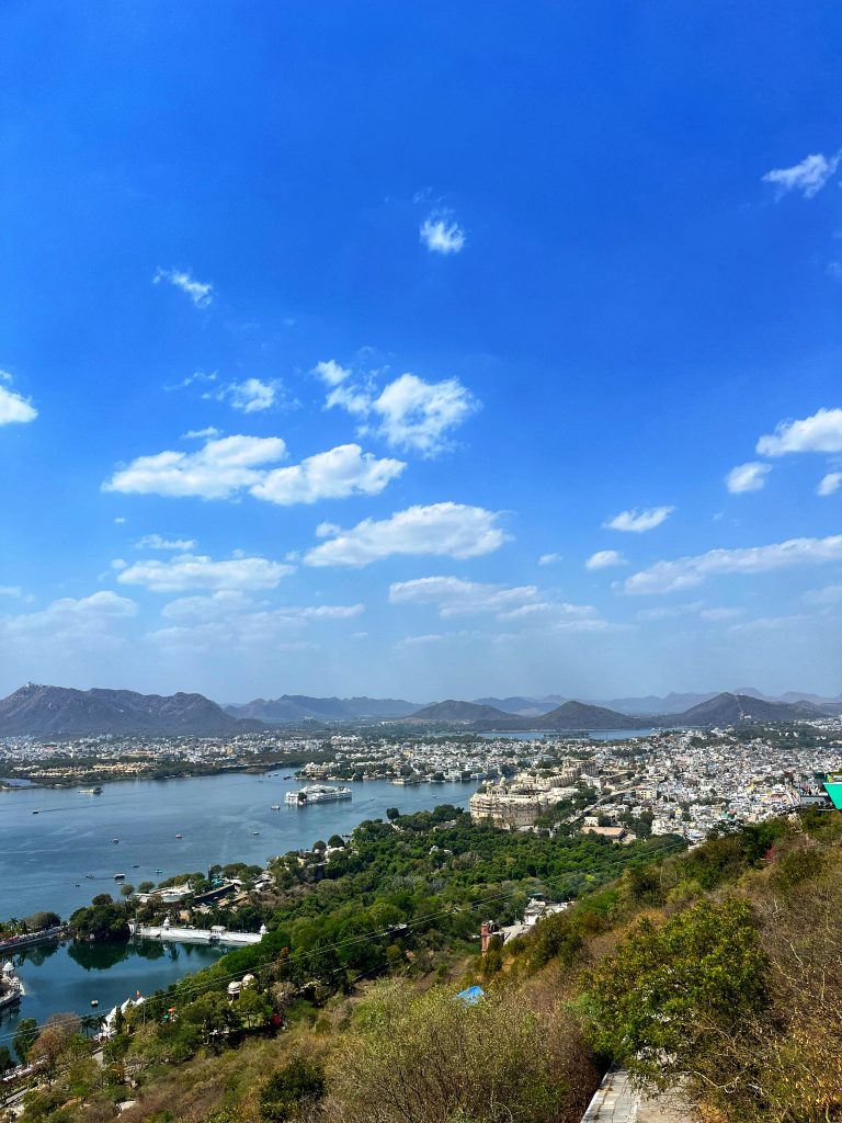 The photo captures a stunning aerial view of a city nestled around a large lake, Udaipur in India. The lake is dotted with small boats and features islands with structures. Surrounding the lake are lush green areas, historic buildings, and a dense cluster of white-washed houses.