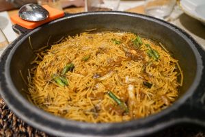 View larger photo: A large black pan filled with Valencian fideuá consisting in cooked vermicelli noodles mixed with vegetables and bits of meat, topped with a sauce. The pan is set on a wooden table with a glass of water and a plate visible in the background.