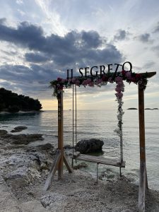  A wooden swing set adorned with flowers and the sign “Il Segreto” stands on a rocky shoreline,  overlooking a calm sea and dramatic
