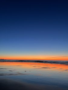 A serene beach at sunset, with a vibrant orange and yellow sky reflecting on the calm water. The horizon separates the warm colors of the sunset from the deep blue sky above.