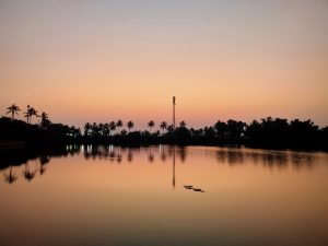 A tranquil lake at sunrise with a silhouette of trees and a communication tower in the background, and the sky transitioning from a soft orange to a light purple hue, all reflected on the calm water surface.