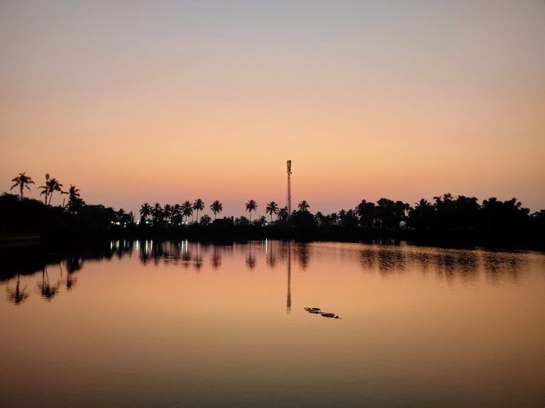 A tranquil lake at sunrise with a silhouette of trees and a communication tower in the background, and the sky transitioning from a soft orange to a light purple hue, all reflected on the calm water surface.