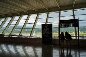 Two people looking at the arrivals panels at Bilbao airport. There's a big windows from where we see two airplanes and the mountains