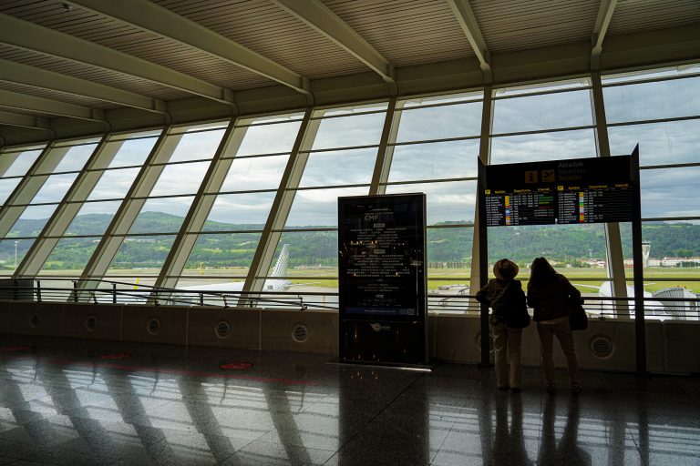 Two people looking at the arrivals panels at Bilbao airport. There’s a big windows from where we see two airplanes and the mountains