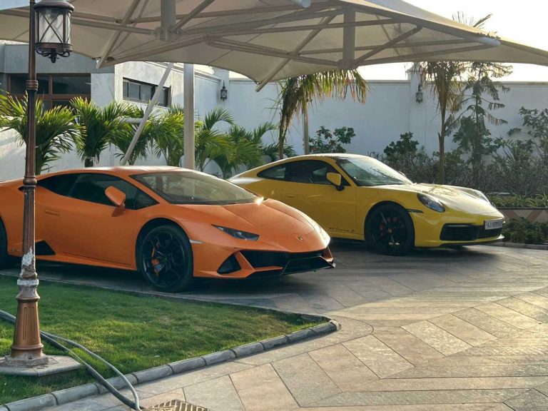 A yellow Porche 911 sports car sitting next to an orange Huracan EVO luxury street racing car under an awning in a courtyard, surrounded by short palm trees