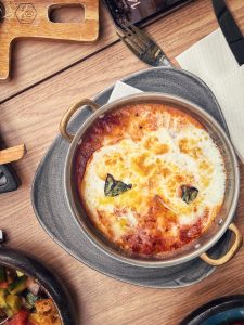 A bubbling dish of baked eggs and tomato sauce garnished with basil, served in a round metal pan on a gray plate.






