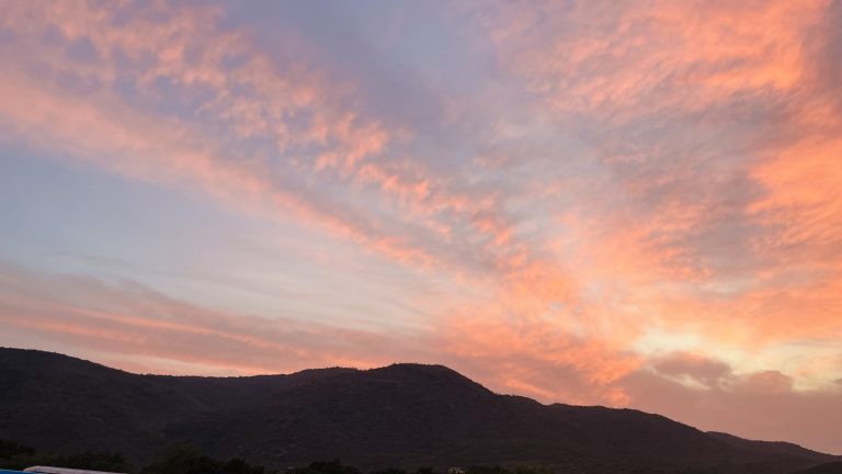 A stunning sunset over a mountainous landscape. The sky is awash with soft hues of pink, orange, and purple, creating a serene and picturesque scene. The mountains are silhouetted against the vibrant sky, with their dark, rolling forms providing a beautiful contrast to the bright, colorful clouds above. The trees at the base of the mountains are ba