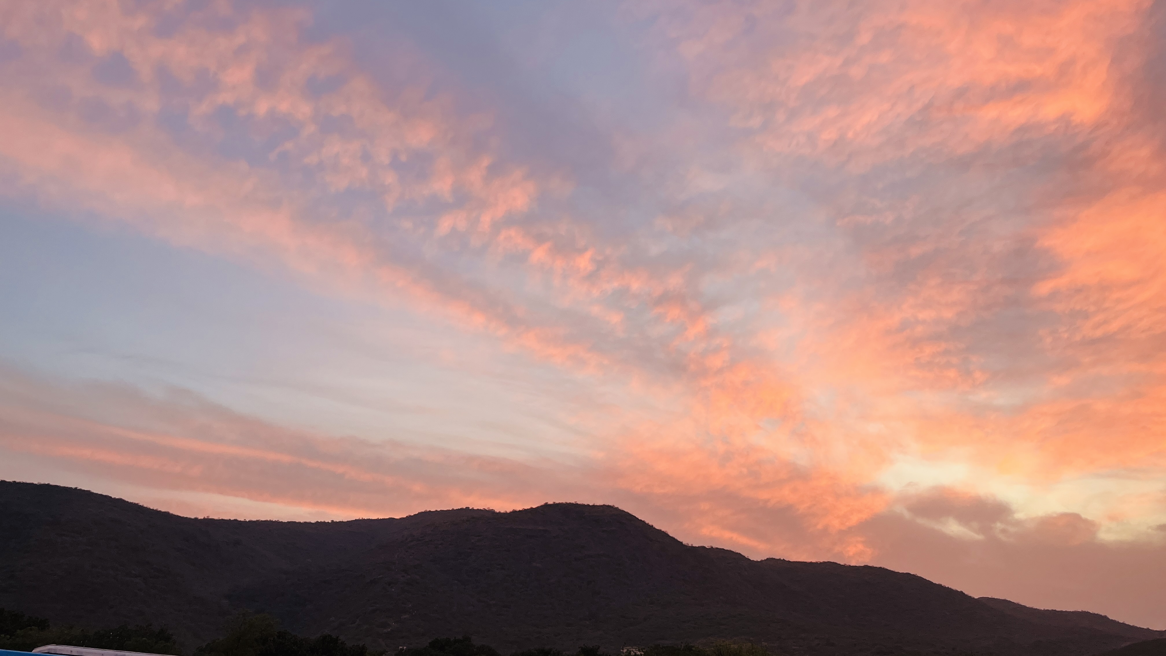 A stunning sunset over a mountainous landscape. The sky is awash with soft hues of pink, orange, and purple, creating a serene and picturesque scene. The mountains are silhouetted against the vibrant sky, with their dark, rolling forms providing a beautiful contrast to the bright, colorful clouds above. The trees at the base of the mountains are ba