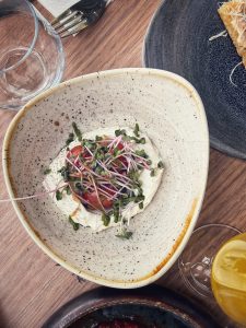 A beautifully plated dish featuring a creamy base topped with sliced tomatoes, fresh greens, and microgreens, set on a rustic ceramic plate.