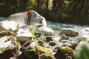 Person standing on a boulder near a flowing river looking out at the scene before them on a sunny day. 