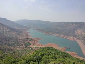 A scenic view of a large reservoir surrounded by hilly terrain. The reservoir has a dam on one side, with a lush green forest in the foreground. The hills have sparse vegetation, showing a mix of brown and green patches. The sky is clear with a few clouds.