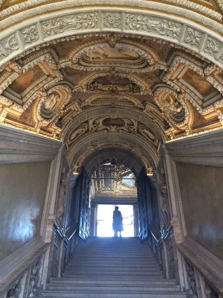 A view up indoor stonework stairs with ornate frescos on the ceiling with a backlit person at the top of the stairs (Venice, Italy)