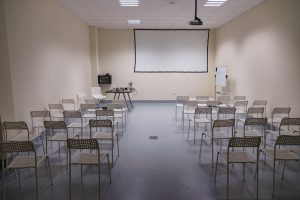 A compact conference room showcasing white chairs, a projector screen, and a whiteboard.