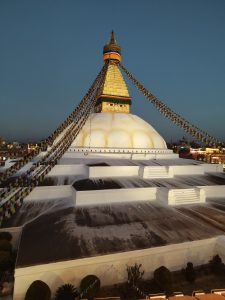 The Boudhanath Stupa at dusk, with its iconic whitewashed dome, colorful prayer flags radiating from the central spire, and the eyes of the Buddha painted on the tower. The surrounding area appears calm with a clear sky above.
