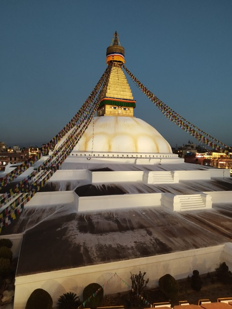 The Boudhanath Stupa at dusk, with its iconic whitewashed dome, colorful prayer flags radiating from the central spire, and the eyes of the Buddha painted on the tower. The surrounding area appears calm with a clear sky above.