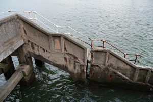 A weathered concrete staircase with rusted metal railings leading down into the water. The structure shows significant signs of erosion and decay.