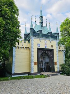 Mirror Maze near to Petrin Park, a medieval-style gatehouse with a yellow and white facade, topped with multiple green spires. The building features a large arched entrance with a metal gate and is adorned with several coats of arms. The structure is surrounded by lush green trees, and a cobblestone pathway leads to the entrance. Prague, Czech Republic.