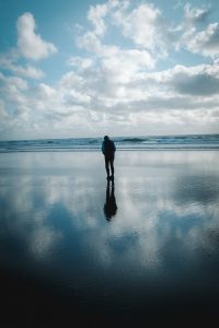 Person on the beach looking down at their feet in the middle of a reflection of the clouds between the sky and the shore. 