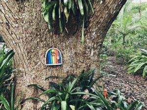 View larger photo: A photo of a fairy door on a live oak tree. The door has rainbow colored wood panels in vertical stripes. There are palmetto bushes around the tree.