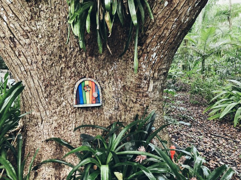 A photo of a fairy door on a live oak tree. The door has rainbow colored wood panels in vertical stripes. There are palmetto bushes around the tree.