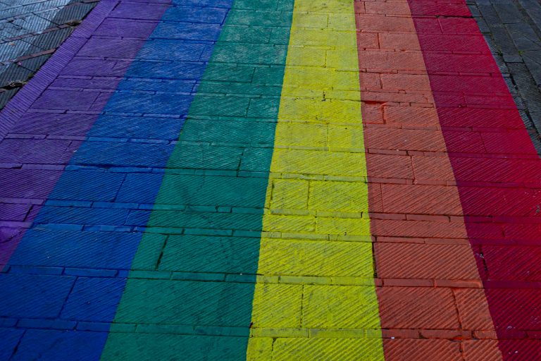 A city street with a rainbow painted on it.  The rainbow stripes run the full length of the image and all are 10s of centimeters wide