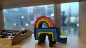 A wooden toy in the shape of a rainbow is placed on a wooden surface with a blurry background of buildings visible through a window. Another wooden tray with small assorted shapes is partially visible on the left side of the image.