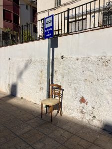 一把带软垫座椅的破旧木椅被放在铺有瓷砖的人行道上，旁边是一个公共汽车站标志。
