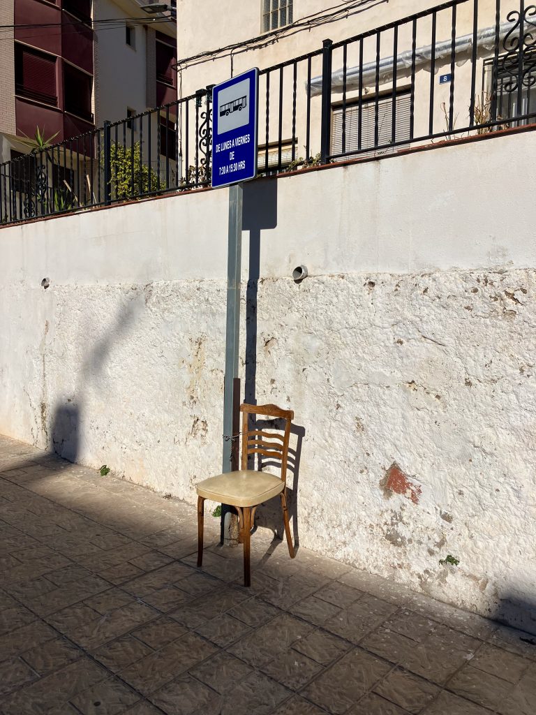 A worn wooden chair with a padded seat is placed on a tiled sidewalk next to a bus stop sign.