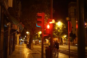 View larger photo: A city street at night with illuminated streetlights, a traffic light displaying a red signal for pedestrians, and a few cyclists riding on the road. The sidewalk and background are dimly lit.