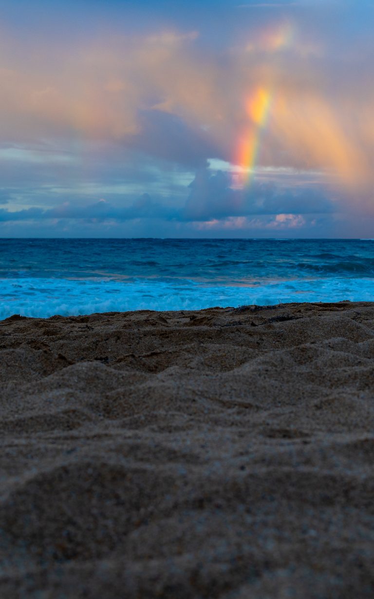 A rainbow above the ocean with a sandy beach in the foreground.