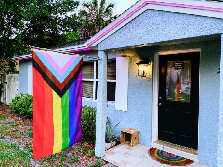 An inclusive pride flag on a pole hanging down in front of a light blue house. The house has pink lights along white trim, a brown door with a sign on it, and a rainbow welcome mat.