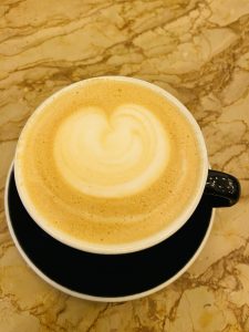 A cup of cappuccino with a heart-shaped foam art on top, placed on a black saucer, resting on a marble table.