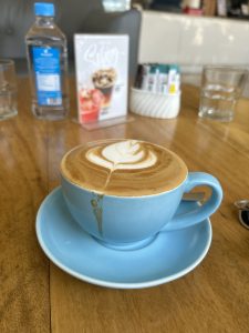 A cup of coffee with a leaf-shaped latte art design on a wooden table, served in a light blue cup and saucer.