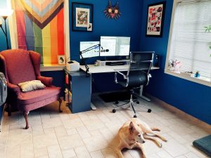 a home office with dark blue walls and a large window. There is a standing desk, an office chair, and an armchair. One wall has a progress pride flag hung up. There is a dog in the foreground sitting on the floor 