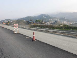 Road construction site with a "20 Speed Limit" sign and traffic cones in the foreground, a partially paved road, construction machinery, piles of gravel, and a hilly landscape with scattered buildings in the background.