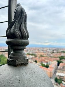  Close-up view of a decorative metal flame-shaped finial at the top of the Mole Antonelliana in Turin, Italy. The blurred background reveals the cityscape with its red-tiled roofs and surrounding buildings under a cloudy sky.