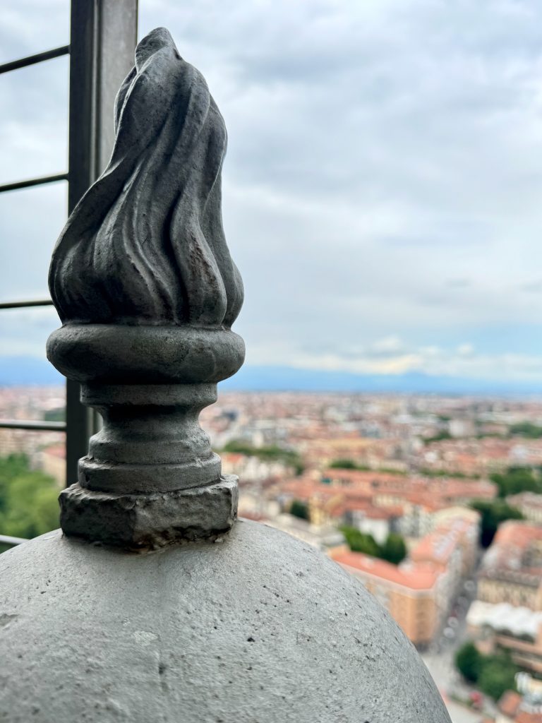 Close-up view of a decorative metal flame-shaped finial at the top of the Mole Antonelliana in Turin, Italy. The blurred background reveals the cityscape with its red-tiled roofs and surrounding buildings under a cloudy sky.