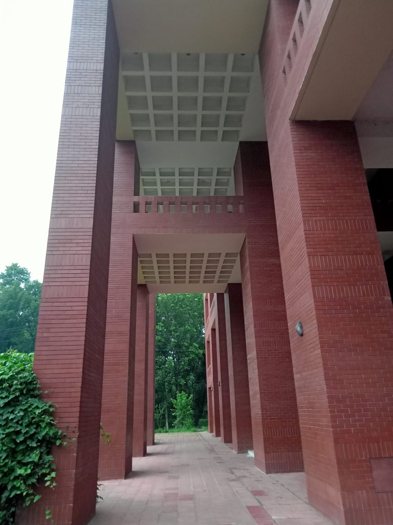 A tall, red brick archway building with a gridded ceiling underpass at Jahangirnagar University. The path below is paved with red bricks and leads to a green, tree-filled area.