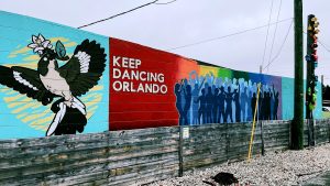 A mural on a brick wall with a lower wood wall in front of it. The mural shows a bird on the left with a flower in its beak. In the middle there is a rainbow gradient, the text "Keep Dancing Orlando", and 49 human outlines to memorialize the 49 victims of the Pulse Club shooting